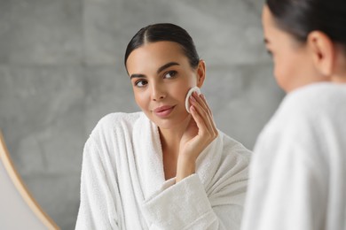 Beautiful woman removing makeup with cotton pad near mirror indoors