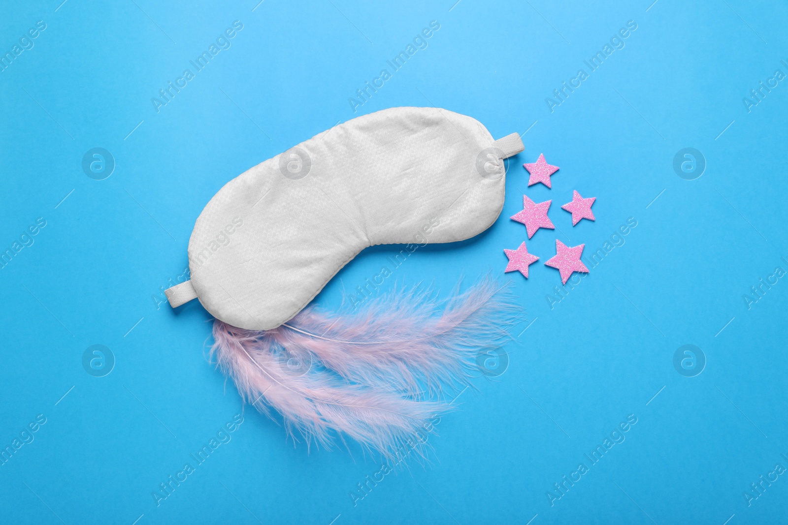 Photo of Soft sleep mask, decorative stars and pink feathers on light blue background, flat lay