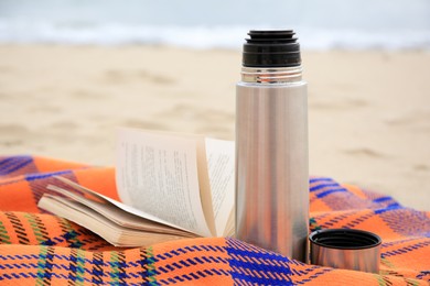 Photo of Metallic thermos with hot drink, open book and plaid on sandy beach near sea