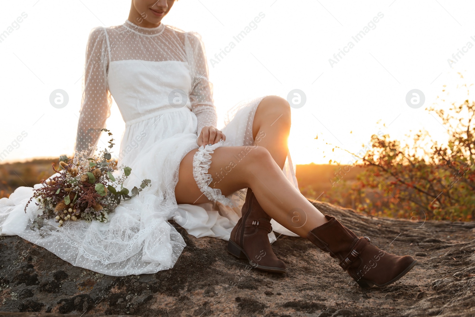 Photo of Beautiful bride with field bouquet sitting on rock at sunset, closeup