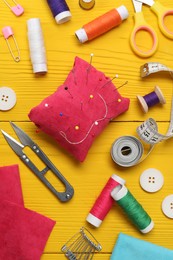Red pincushion with pins and other sewing tools on yellow wooden table, flat lay