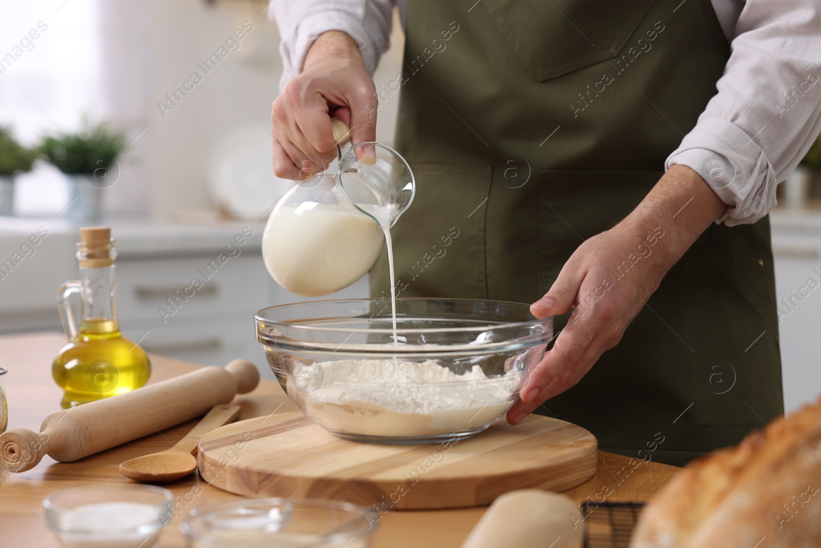 Photo of Making bread. Man pouring milk into bowl with flour at wooden table in kitchen, closeup