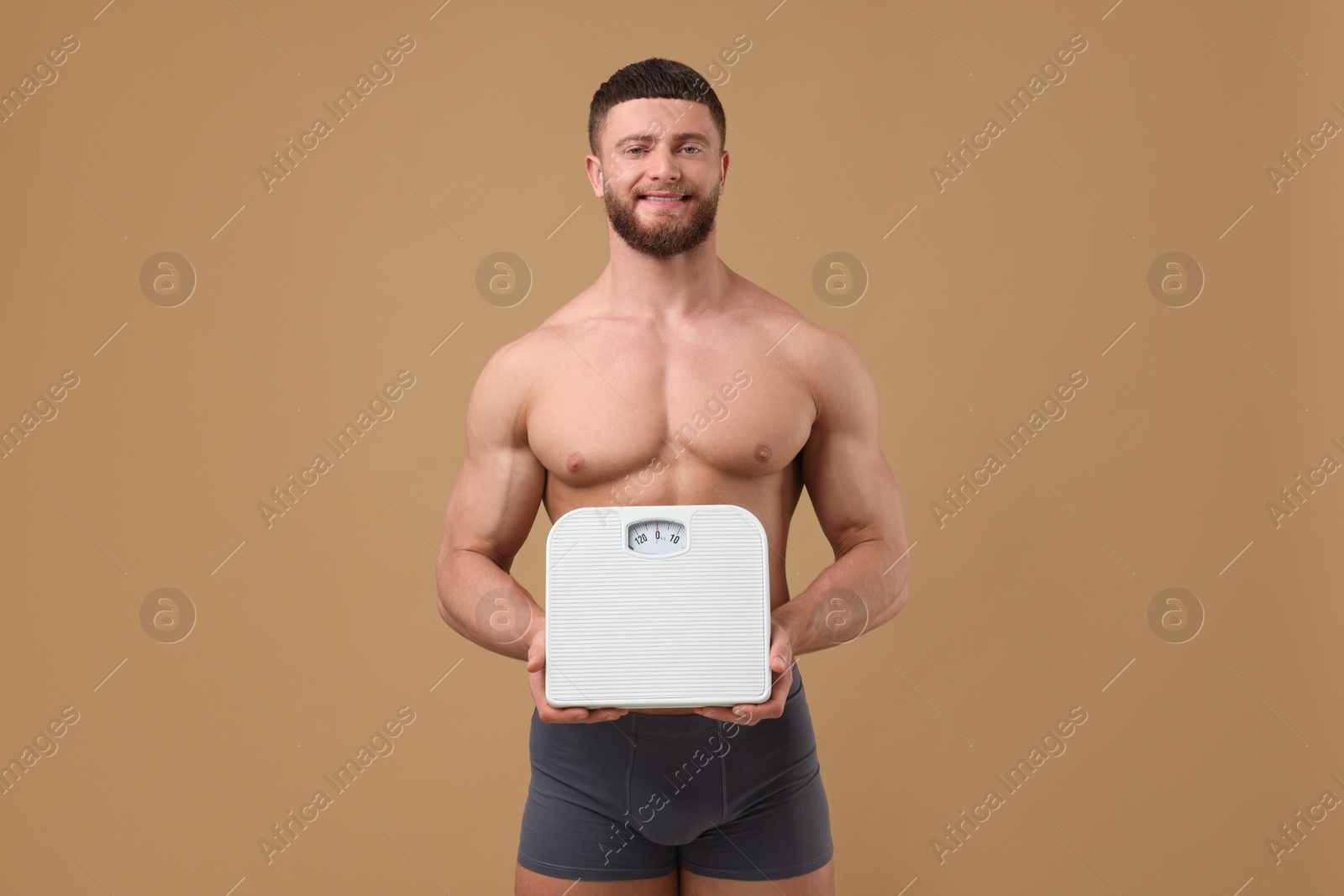 Photo of Smiling athletic man holding scales on brown background. Weight loss concept