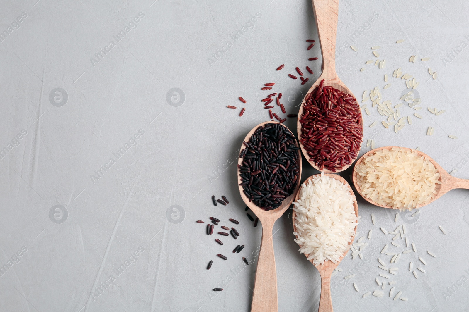 Photo of Flat lay composition with brown and other types of rice in wooden spoons on grey background. Space for text