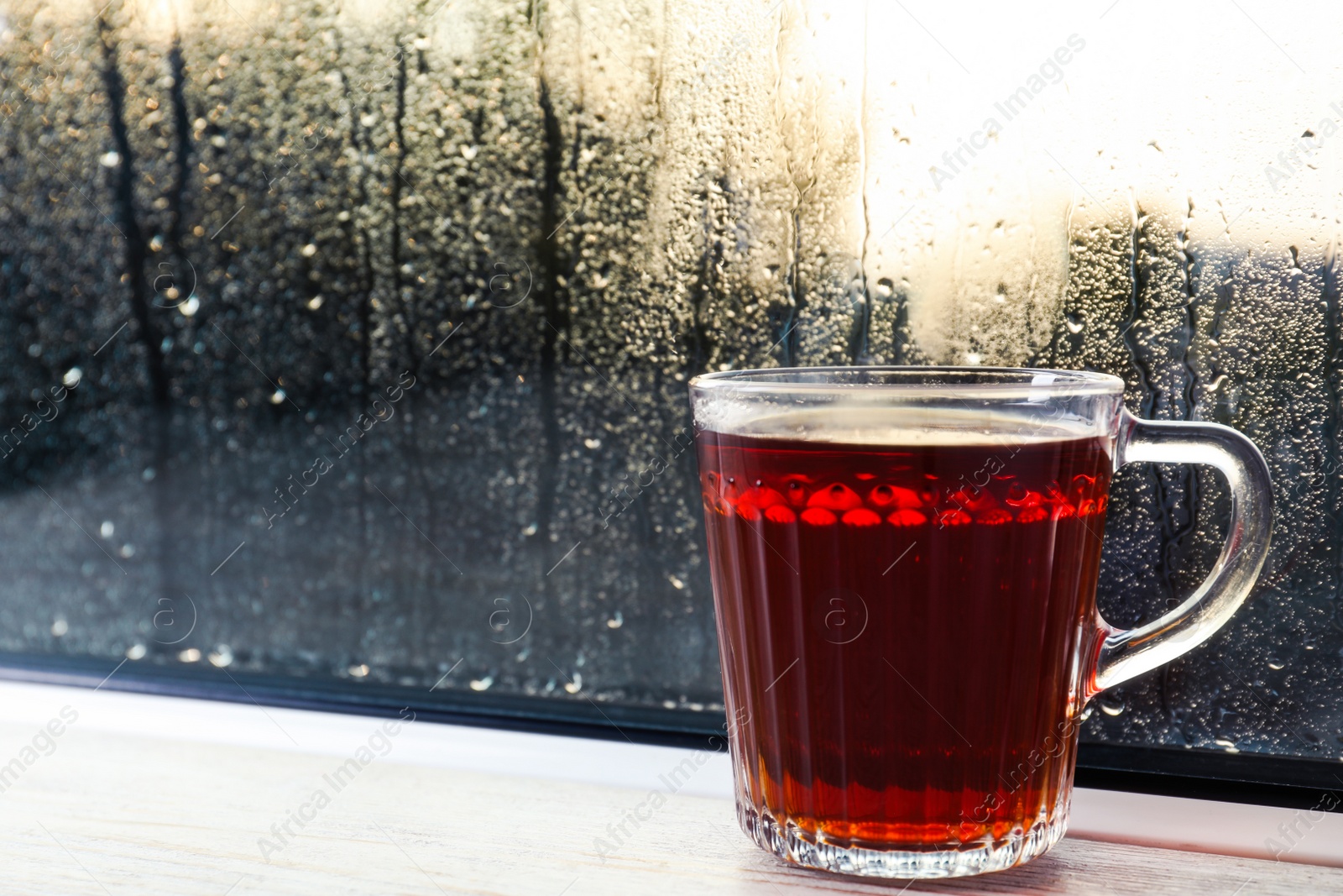 Photo of Glass mug of hot tea on white wooden window sill, closeup. Rainy weather