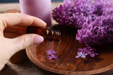 Woman pouring lilac essential oil into bowl at wooden table, closeup