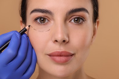 Doctor drawing marks on woman's face for cosmetic surgery operation against beige background, closeup