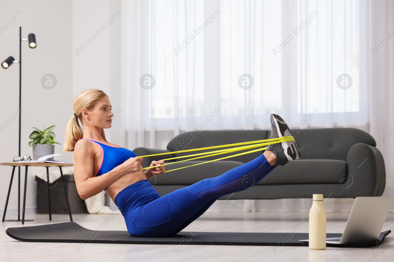 Photo of Fit woman doing exercise with fitness elastic band near laptop on mat at home