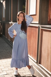 Beautiful young woman in stylish blue dress with handbag near wooden building outdoors