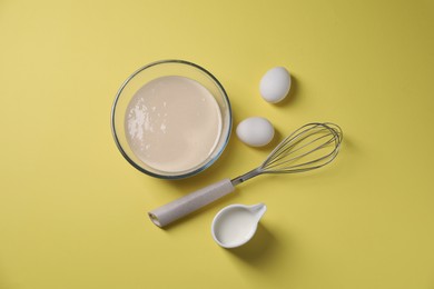 Photo of Metal whisk, dough in bowl, eggs and milk on pale yellow background, flat lay