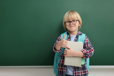 Happy little school child with notebooks showing thumbs up near chalkboard. Space for text