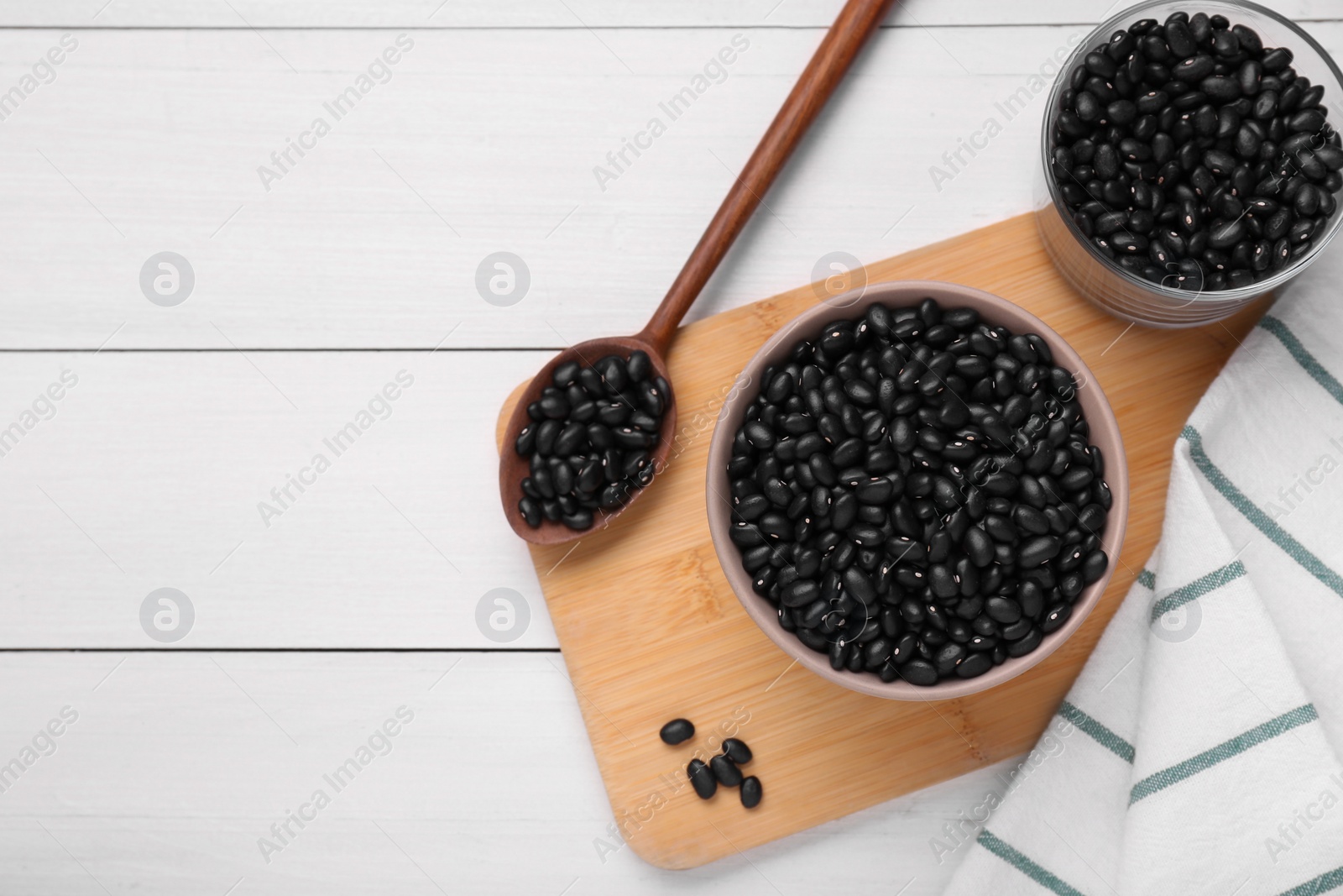 Photo of Bowl and spoon of raw black beans on white wooden table, flat lay. Space for text