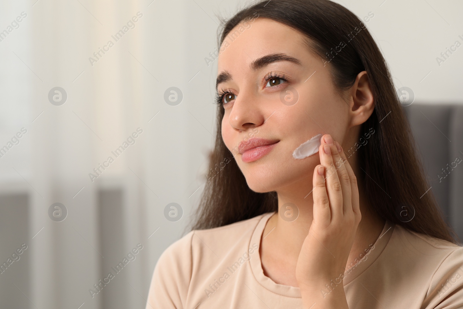 Photo of Young woman with dry skin applying cream onto her face indoors. Space for text