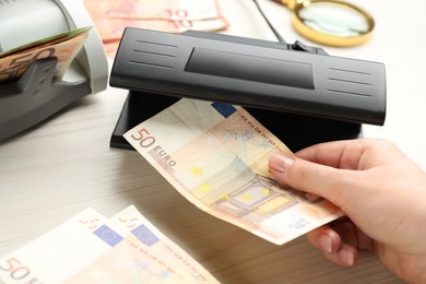 Photo of Woman checking Euro banknote with currency detector at white wooden table, closeup. Money examination device
