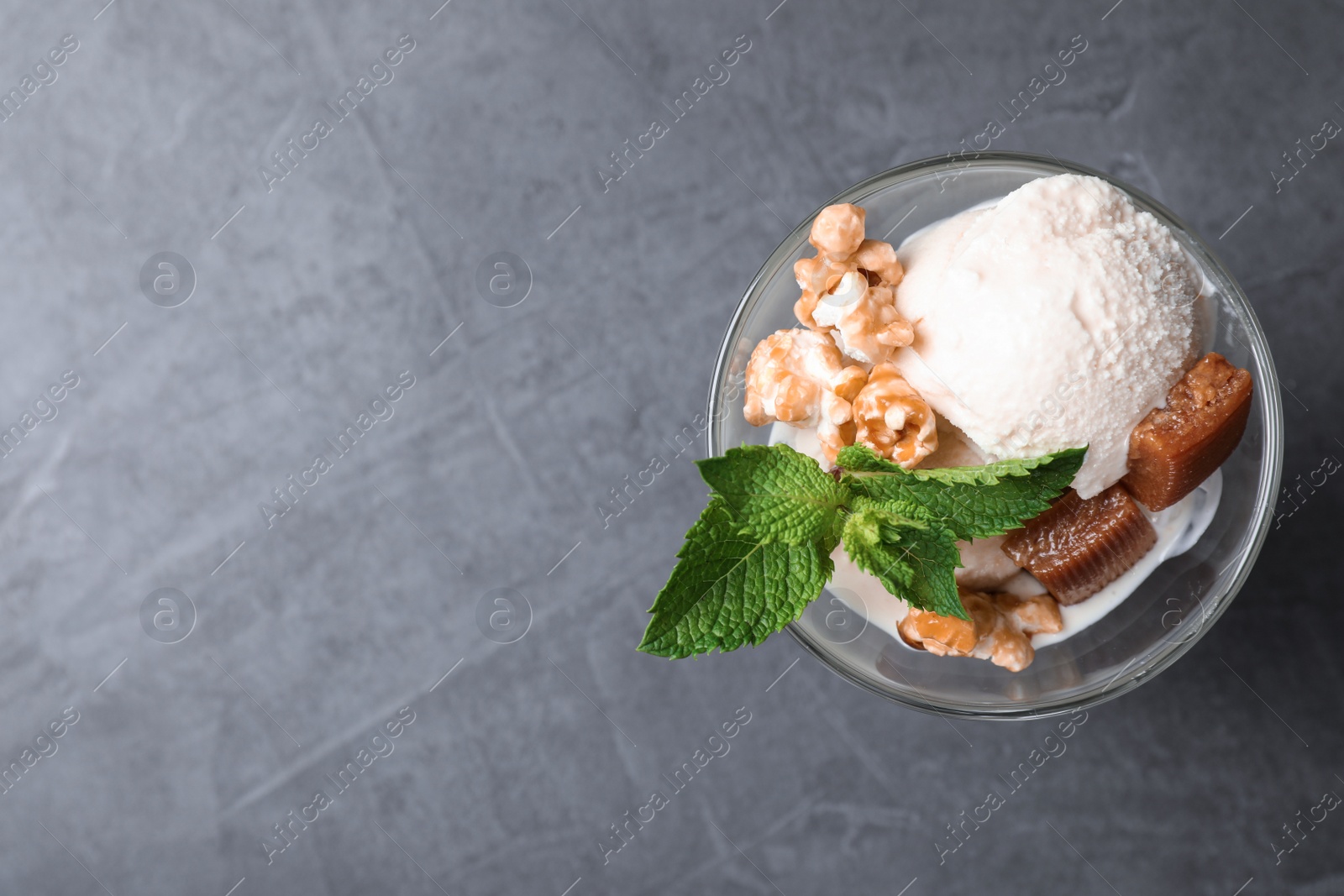 Photo of Glass bowl of ice cream with caramel candies, mint and popcorn on grey table, top view. Space for text