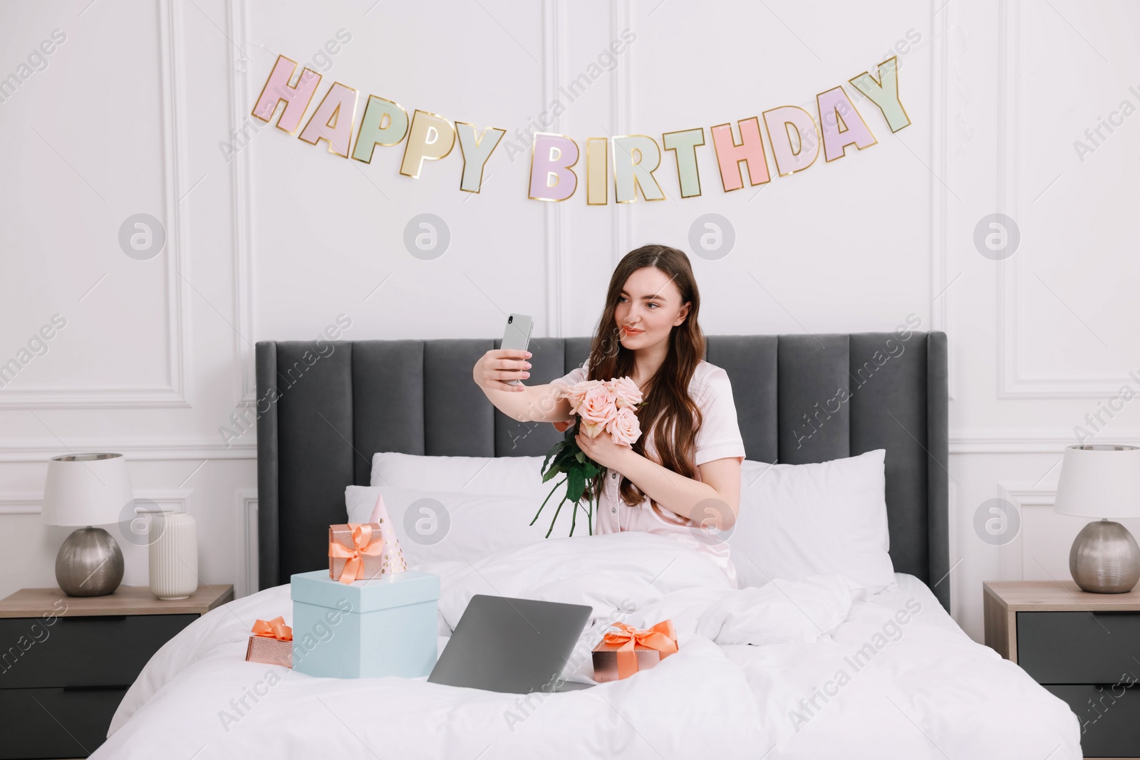 Photo of Beautiful young woman taking selfie with rose flowers, laptop and gift boxes on bed in room. Happy Birthday