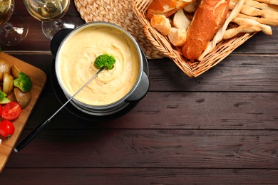 Fork with piece of broccoli, melted cheese in fondue pot, wine and snacks on wooden table, flat lay. Space for text