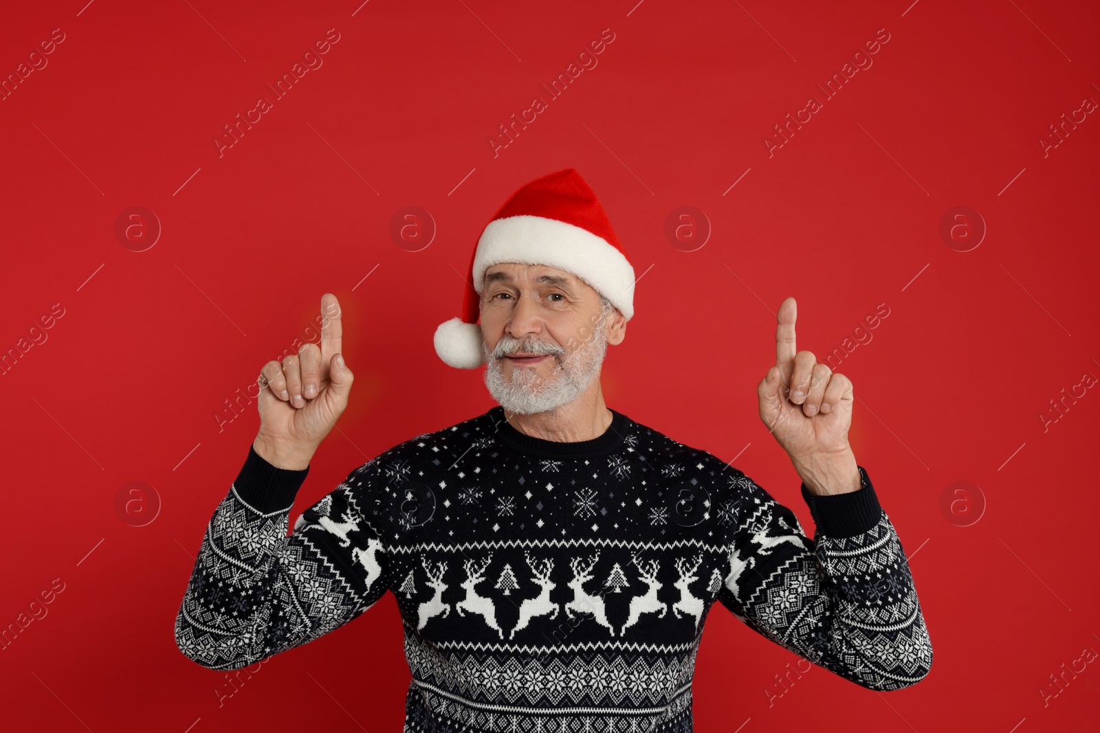 Photo of Senior man in Christmas sweater and Santa hat pointing at something on red background