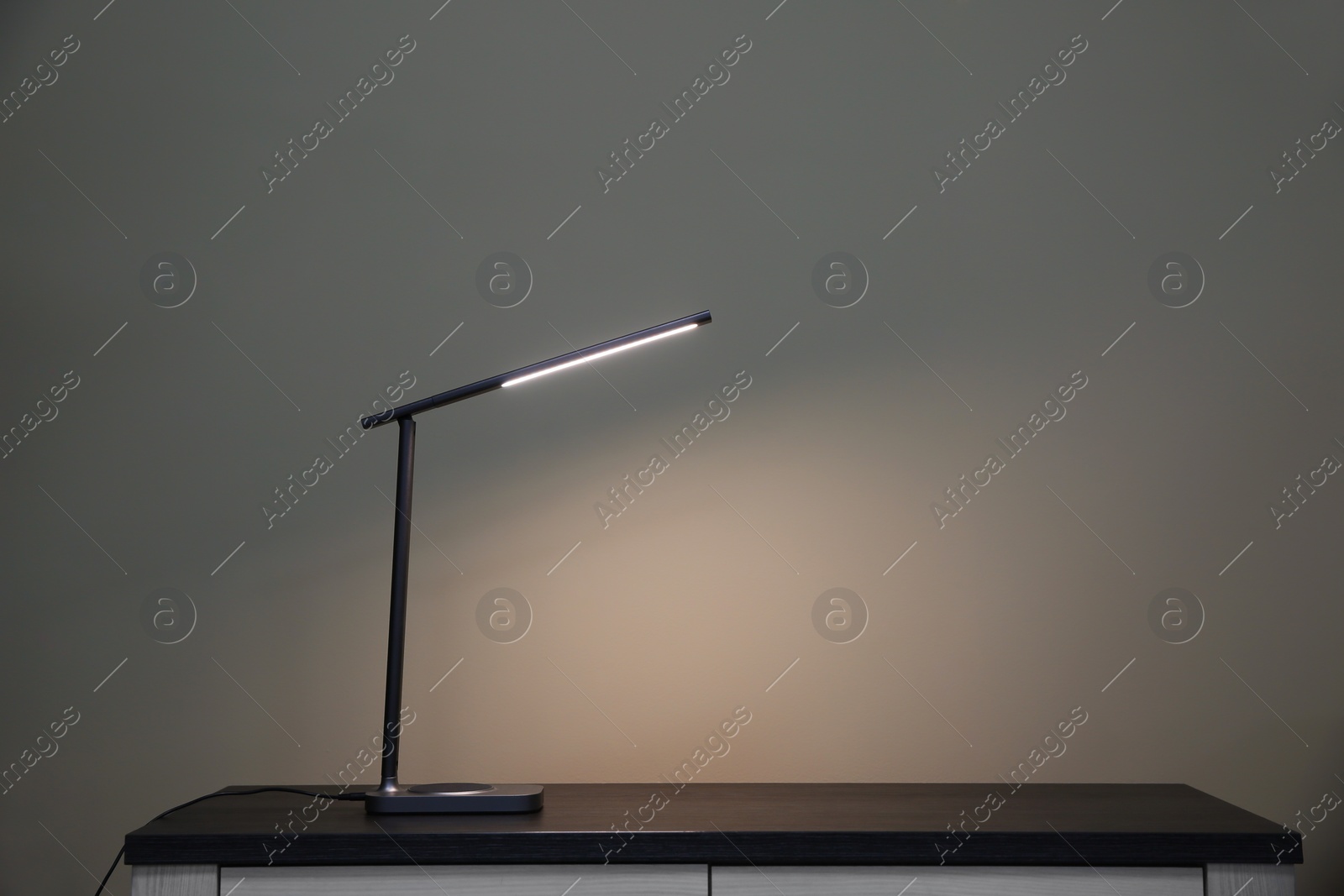 Photo of Wooden table with modern lamp near light grey wall, space for text. Interior element