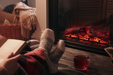 Woman with cup of tea reading book near fireplace at home, closeup