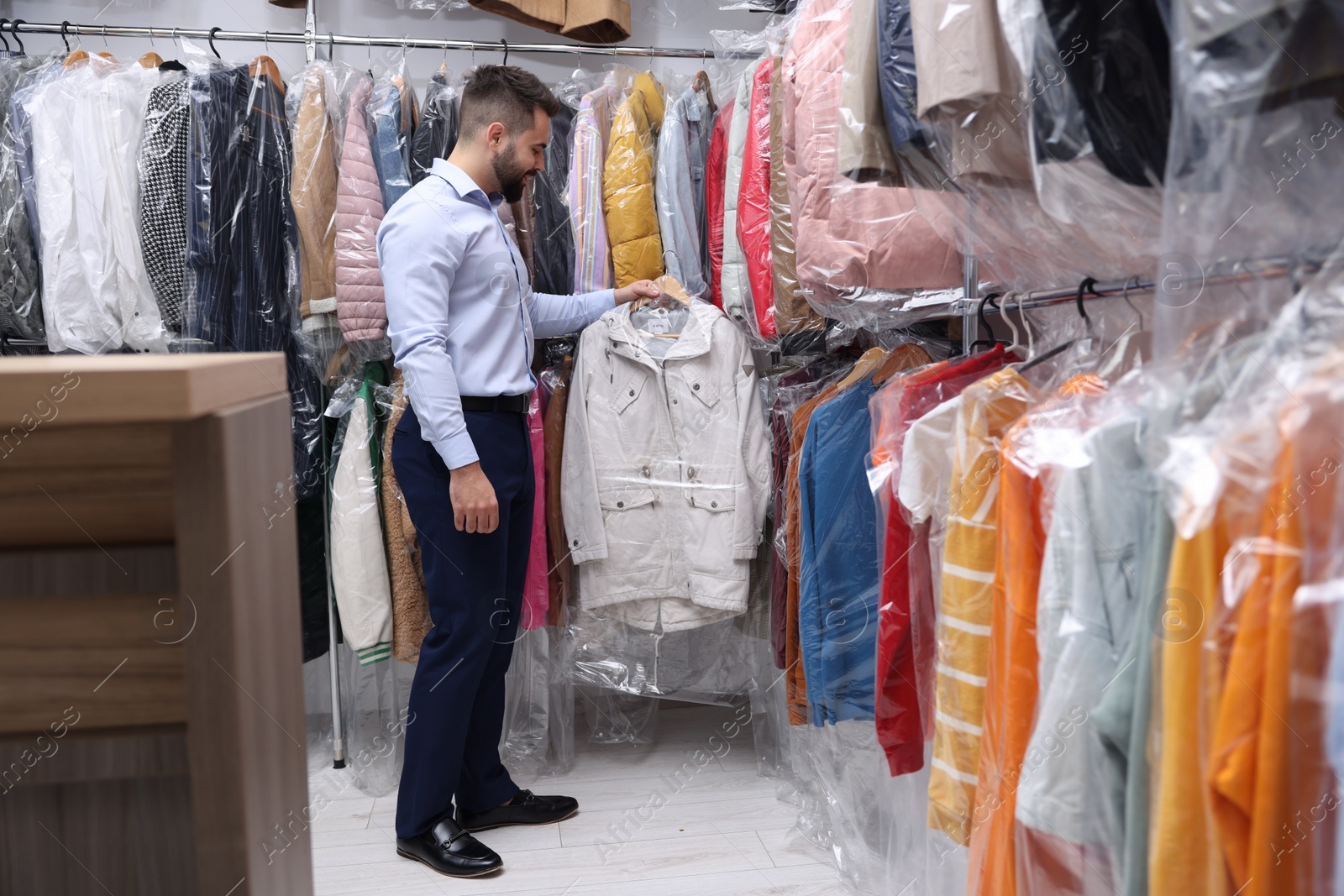 Photo of Dry-cleaning service. Happy worker holding hanger with jacket in plastic bag near other clothes indoors