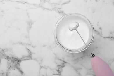 Mini mixer (milk frother) and whipped milk in glass on white marble table, top view. Space for text