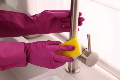 Photo of Woman in gloves cleaning faucet of kitchen sink with sponge, closeup