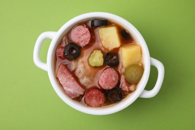 Photo of Meat solyanka soup with thin dry smoked sausages in pot on light green background, top view