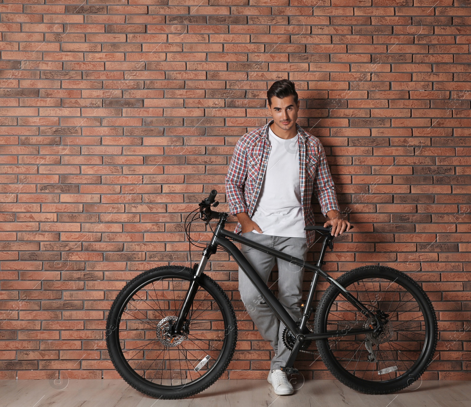 Photo of Handsome young man with modern bicycle near brick wall indoors