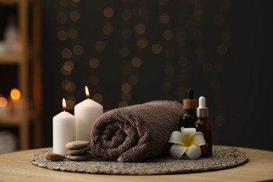Photo of Spa composition. Rolled towel, cosmetic products, stones, burning candles and plumeria flower on table indoors. Space for text