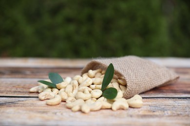 Tasty cashew nuts, green leaves and burlap bag on wooden table outdoors, space for text