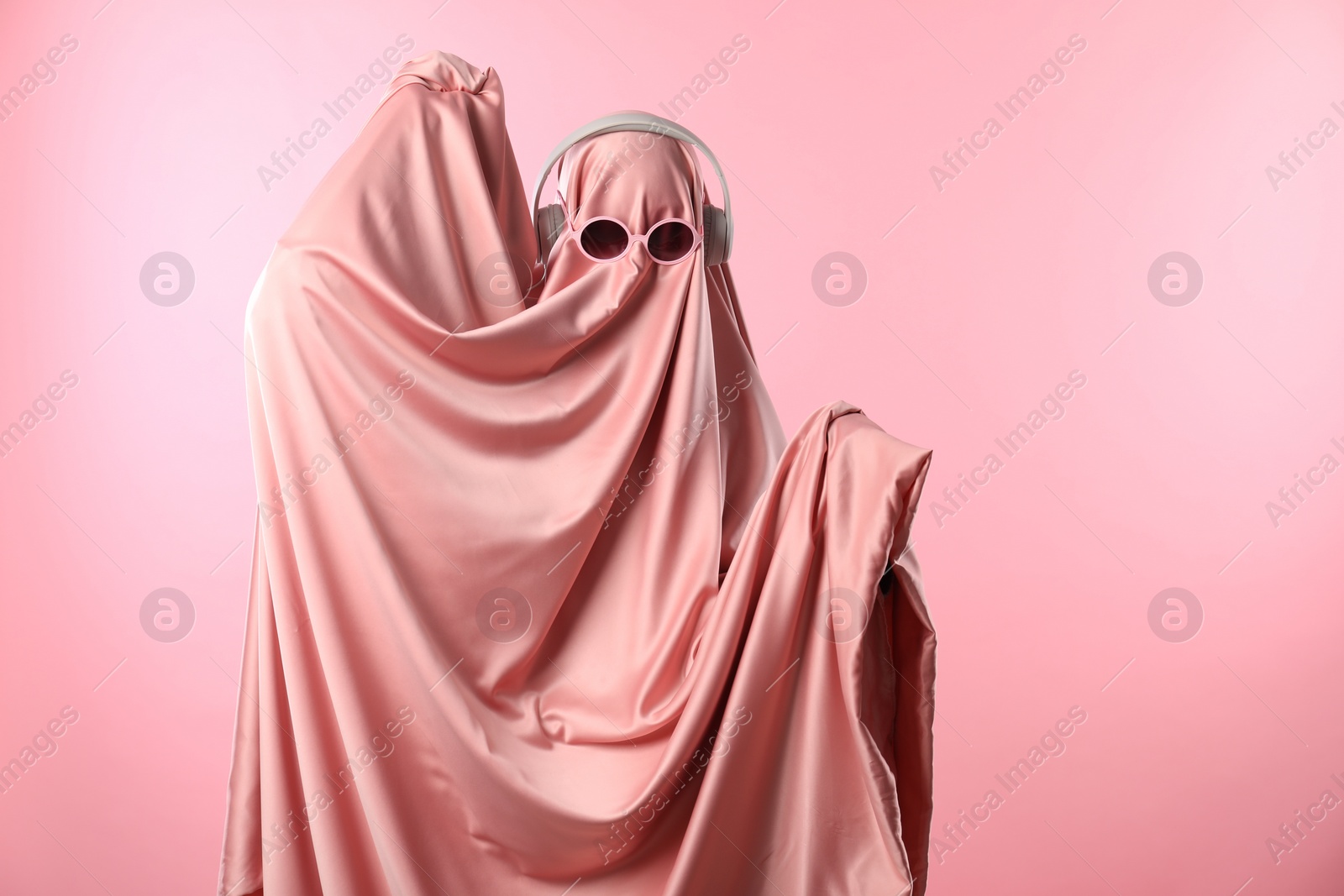 Photo of Glamorous ghost. Woman in sheet with sunglasses and headphones on pink background, space for text