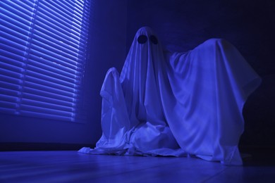 Photo of Creepy ghost. Woman covered with sheet near window in blue light, low angle view. Space for text