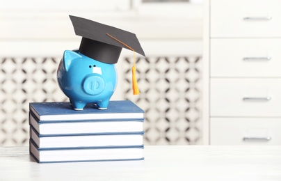 Piggy bank with graduation hat and books on table against blurred background. Space for text