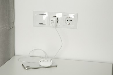 Photo of Charging case of modern wireless earphones plugged into power socket and smartphone on white table indoors