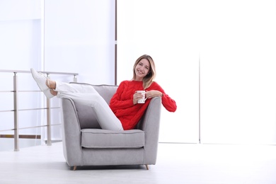 Attractive young woman with cup of tea in armchair indoors. Space for text
