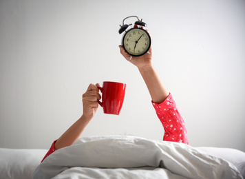 Photo of Woman with cup and alarm clock lying in bed, closeup. Morning time
