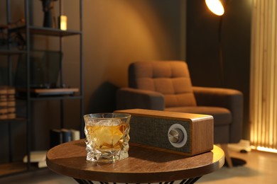 Photo of Glass of whiskey and portable bluetooth speaker on wooden table in room. Relax at home