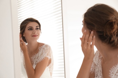 Young bride in beautiful wedding dress near mirror indoors