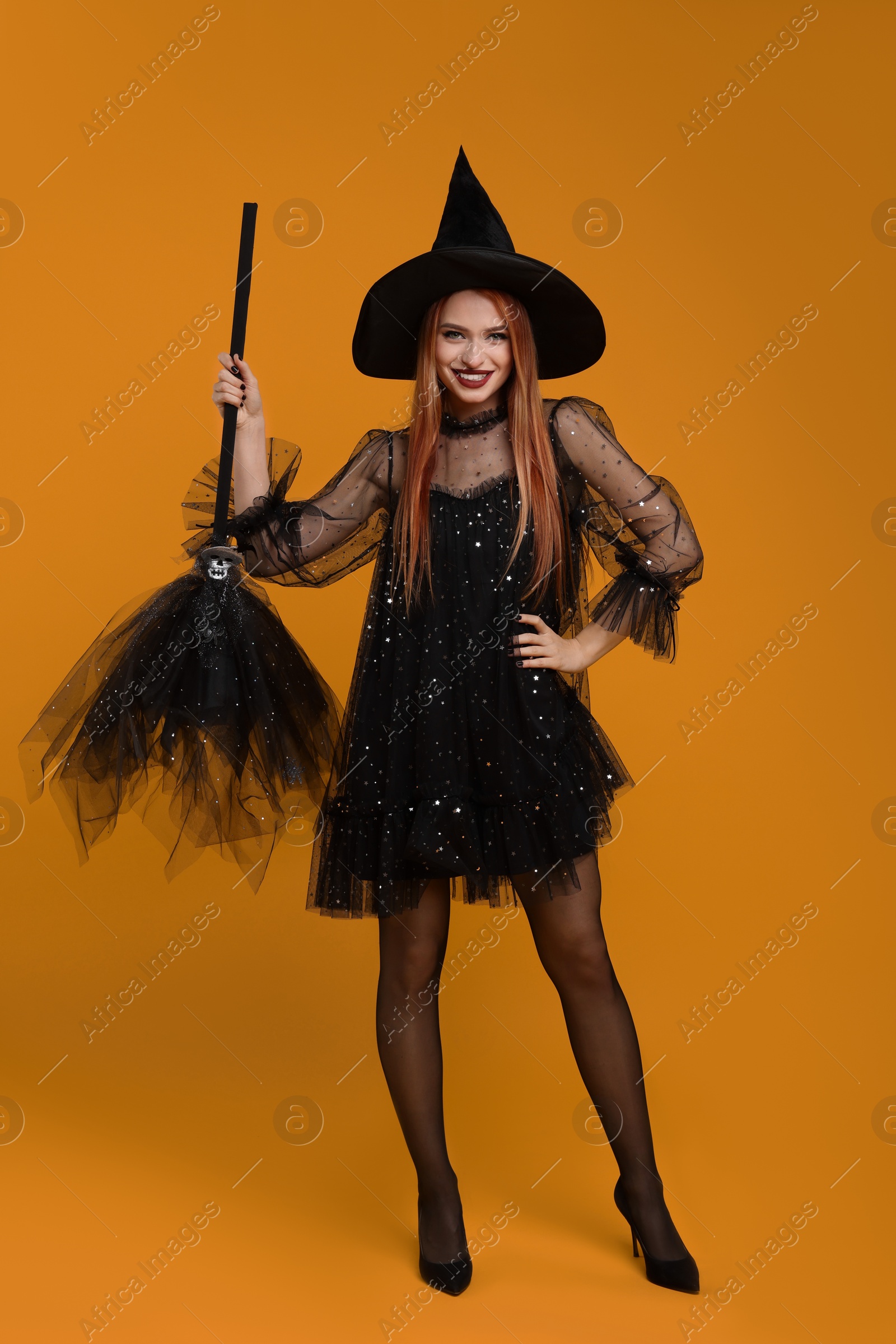 Photo of Happy young woman in scary witch costume with broom on orange background. Halloween celebration