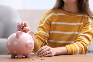 Photo of Woman putting coin into piggy bank at wooden table, closeup