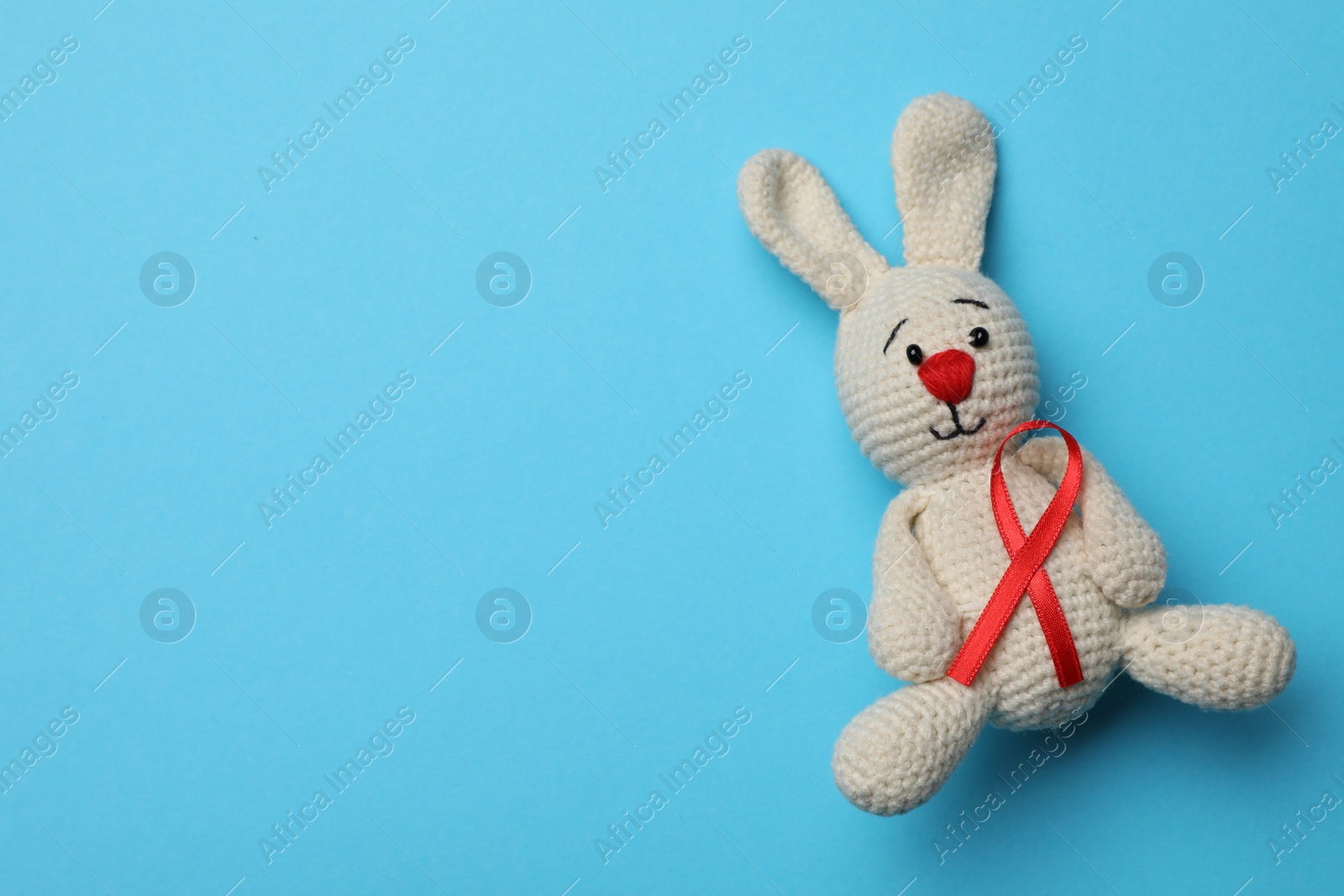 Photo of Cute knitted toy bunny with red ribbon on blue background, top view and space for text. AIDS disease awareness