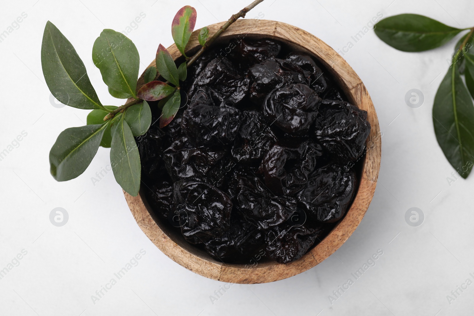 Photo of Sweet dried prunes in bowl and green leaves on white table, top view