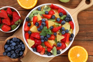 Delicious fresh fruit salad in bowl and ingredients on wooden table, flat lay