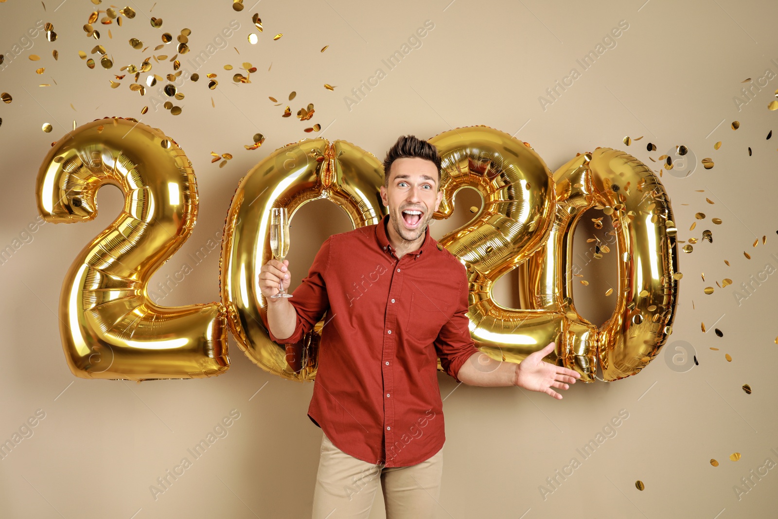 Photo of Excited young man with glass of champagne near golden 2020 balloons on beige background. New Year celebration