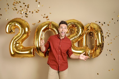 Excited young man with glass of champagne near golden 2020 balloons on beige background. New Year celebration