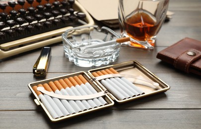 Photo of Open case with tobacco filter cigarettes, ashtray, wallet and alcohol drink near vintage typewriter on wooden table