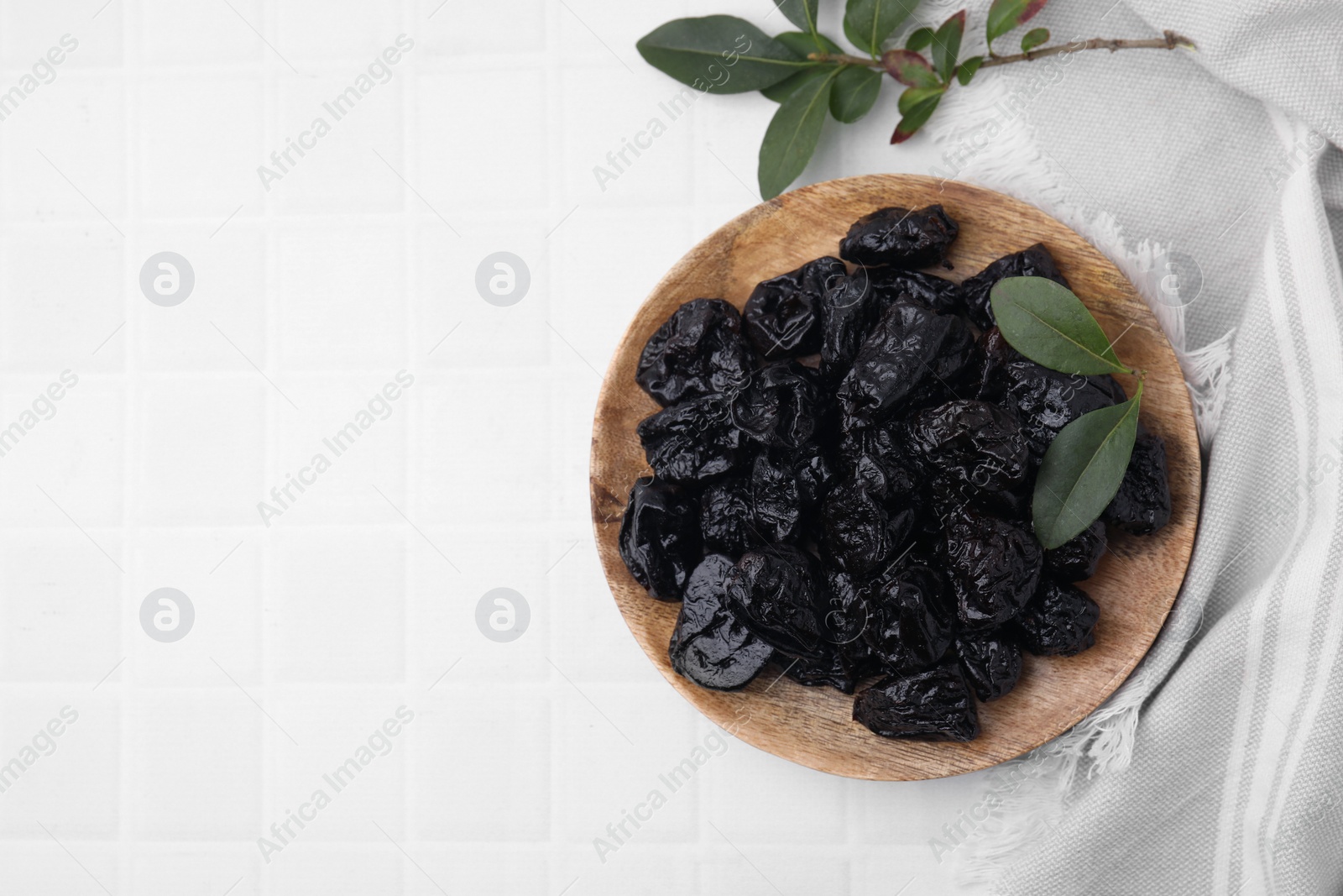 Photo of Sweet dried prunes and green leaves on white tiled table, top view. Space for text