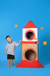 Cute little boy playing with cardboard rocket on light blue background