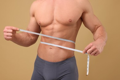 Athletic man measuring waist with tape on brown background, closeup. Weight loss concept